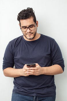 Young man in casual outfit using smartphone indoors, leaning against a white wall.